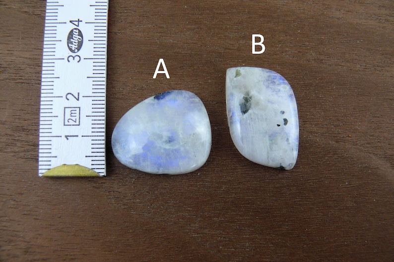 Puede incluir: Dos piedras de luna pulidas, etiquetadas A y B, con una regla para la escala. La piedra A tiene forma ovalada, mientras que B tiene una forma m&aacute;s irregular. Ambas piedras tienen una base blanca lechosa con inclusiones azules y negras.