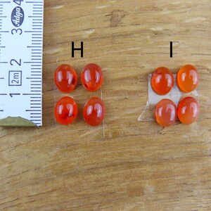 Op de afbeelding: Zes ovale, doorschijnende, oranjerode edelstenen worden getoond. Een liniaal met centimeter markeringen staat naast de edelstenen. De edelstenen liggen op een houten oppervlak, met twee sets van drie edelstenen.