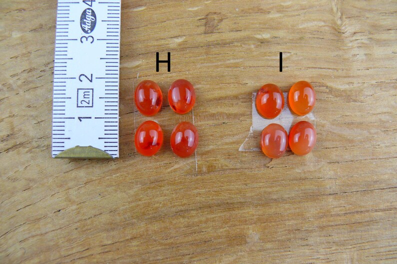 Op de afbeelding: Zes ovale, doorschijnende, oranjerode edelstenen worden getoond. Een liniaal met centimeter markeringen staat naast de edelstenen. De edelstenen liggen op een houten oppervlak, met twee sets van drie edelstenen.