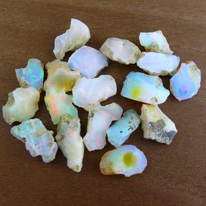 Könnte beinhalten: Eine Sammlung roher Opal-Edelsteine, die eine Vielzahl von Farben wie Weiß, Blau, Gelb und Grün aufweisen. Die Opale haben eine raue, natürliche Textur und liegen auf einer braunen Holzoberfläche.