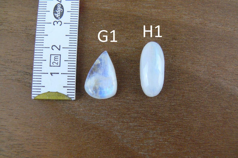 May include: Two polished moonstone cabochons labeled G1 and H1 next to a white measuring tape. The G1 stone is pear-shaped with blue iridescence, while the H1 stone is oval-shaped and white. The measuring tape shows a scale in centimeters.