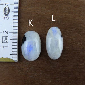 Puede incluir: Dos cabujones ovalados de piedra lunar con una regla para la escala. Las piedras son blancas con inclusiones azules y negras. La regla muestra las medidas en cent&iacute;metros. Las piedras est&aacute;n sobre una superficie de madera marr&oacute;n.