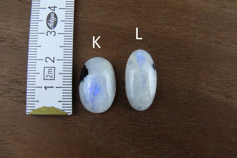 Puede incluir: Dos cabujones ovalados de piedra lunar con una regla para la escala. Las piedras son blancas con inclusiones azules y negras. La regla muestra las medidas en cent&iacute;metros. Las piedras est&aacute;n sobre una superficie de madera marr&oacute;n.