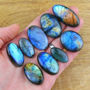 May include: A selection of polished labradorite gemstones in various oval shapes. The stones exhibit iridescent flashes of blue, green, and gold. The gemstones are held in a hand, highlighting their natural beauty and unique patterns. The background is a light brown wooden surface.