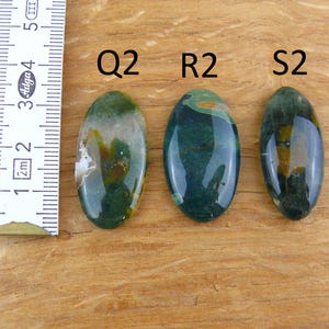 Op de afbeelding: Drie ovaalvormige, gepolijste groene en gele edelstenen met de labels Q2, R2 en S2. Een wit meetlint is aan de linkerkant geplaatst en laat zien dat de stenen ongeveer 4 cm lang zijn. De stenen hebben een gladde, glanzende afwerking.