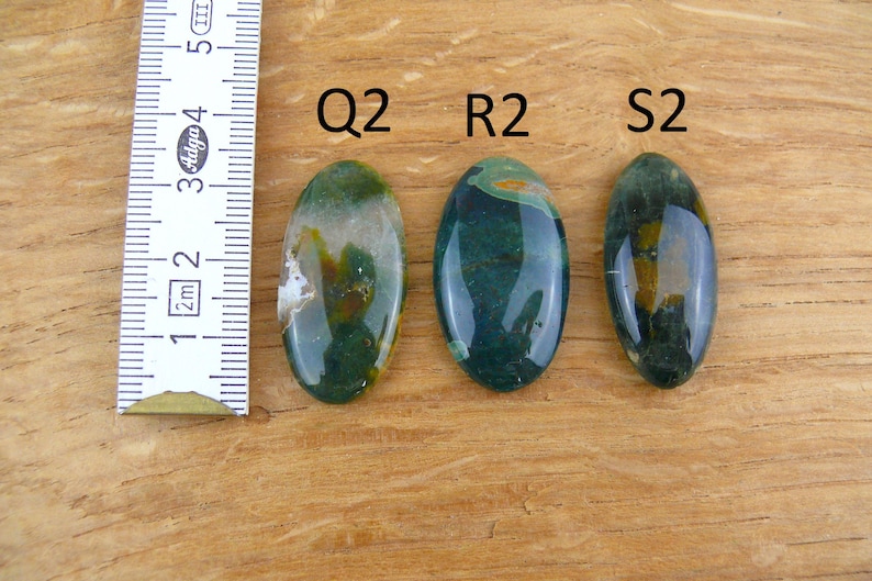 Op de afbeelding: Drie ovaalvormige, gepolijste groene en gele edelstenen met de labels Q2, R2 en S2. Een wit meetlint is aan de linkerkant geplaatst en laat zien dat de stenen ongeveer 4 cm lang zijn. De stenen hebben een gladde, glanzende afwerking.