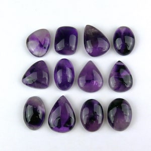 Könnte beinhalten: Eine Sammlung von zwölf polierten Amethyst-Edelsteinen, die in drei Reihen angeordnet sind. Die Steine haben verschiedene Formen, darunter Ovale, Tropfen und abgerundete Rechtecke, und zeigen tiefe violette Farbtöne mit Farbvariationen.
