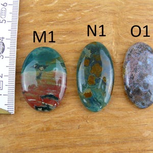 Op de afbeelding: Drie ovaalvormige gepolijste edelstenen met de labels M1, N1 en O1. M1 heeft rode, groene en zwarte patronen. N1 toont groen met bruine vlekken. O1 vertoont gevlekte grijze en blauwe tinten. Een liniaal toont een lengte van 3,2 cm.