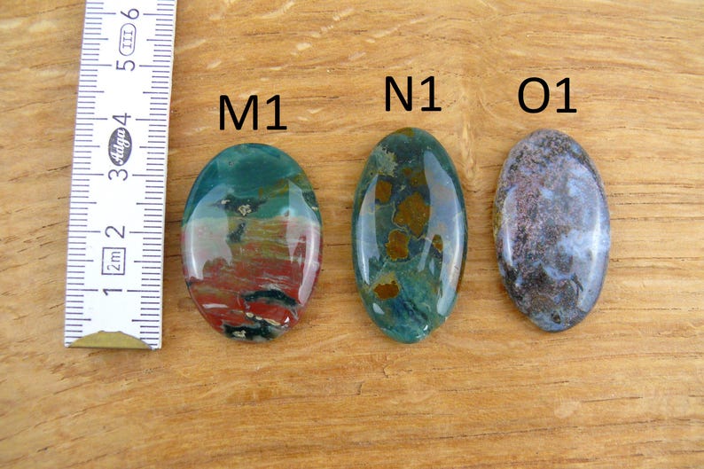 Op de afbeelding: Drie ovaalvormige gepolijste edelstenen met de labels M1, N1 en O1. M1 heeft rode, groene en zwarte patronen. N1 toont groen met bruine vlekken. O1 vertoont gevlekte grijze en blauwe tinten. Een liniaal toont een lengte van 3,2 cm.