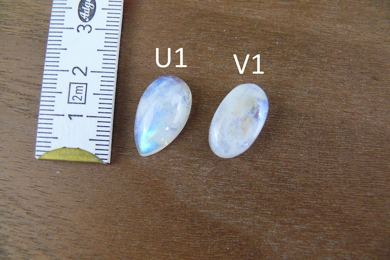 May include: Two polished moonstone cabochons labeled U1 and V1, with U1 being pear-shaped and V1 being oval. A white measuring tape is positioned to the left, showing the stones are approximately 1.25 inches in length for the larger piece.