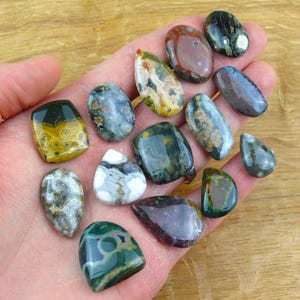 May include: A collection of fifteen polished, multi-colored cabochon gemstones in various shapes including teardrops, ovals, and rectangles. The stones feature diverse patterns and hues, including shades of green, yellow, brown, white, and grey, with smooth, glossy surfaces reflecting light.