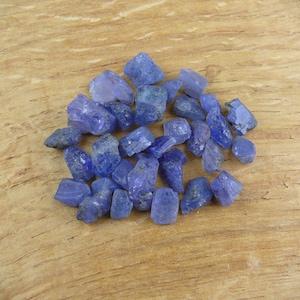Pode incluir: Uma coleção de pedras preciosas de tanzanita brutas, não cortadas, em tons de azul e roxo. As pedras preciosas estão espalhadas sobre uma superfície de madeira marrom clara, mostrando suas estruturas cristalinas naturais.