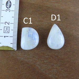 May include: Two polished moonstone cabochons labeled C1 and D1, positioned next to a white measuring tape. The C1 stone is semi-circular, while the D1 stone is teardrop-shaped. Both stones exhibit a translucent white appearance with blue sheen.