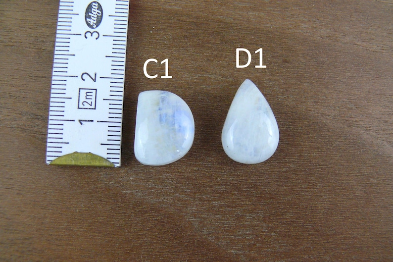 May include: Two polished moonstone cabochons labeled C1 and D1, positioned next to a white measuring tape. The C1 stone is semi-circular, while the D1 stone is teardrop-shaped. Both stones exhibit a translucent white appearance with blue sheen.