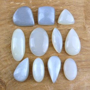 Puede incluir: Colección de piedras preciosas pulidas en diversas formas, incluyendo ovaladas, en forma de lágrima y cuadradas. Las piedras tienen un color blanco lechoso y gris pálido nacarado, con una superficie lisa y reflectante. Las piedras preciosas están dispuestas sobre una superficie de madera.