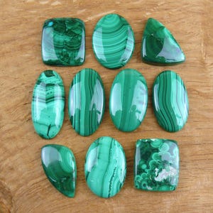 Op de afbeelding: Een assortiment gepolijste malachiet edelstenen in diverse vormen, waaronder ovaal, rechthoekig en driehoekig. De stenen vertonen een levendige groene kleur met kenmerkende bandpatronen. De edelstenen liggen op een houten oppervlak.