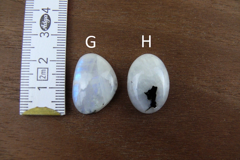 Puede incluir: Dos cabujones de piedra lunar pulidos, uno irregular y otro ovalado, con una regla para la escala. Las piedras son blancas con destellos azules iridiscentes. La piedra ovalada tiene una inclusi&oacute;n negra. El fondo es una superficie de madera oscura.
