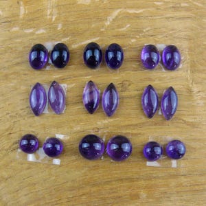 May include: An assortment of polished amethyst gemstones in various shapes, including oval, marquise, and round. The gemstones are a deep purple color and are arranged on a wooden surface.