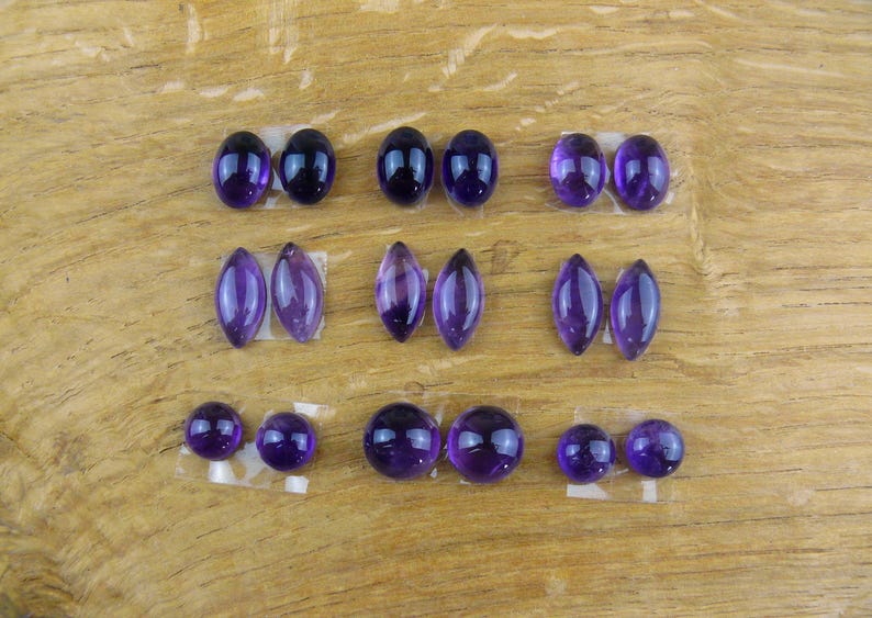 May include: An assortment of polished amethyst gemstones in various shapes, including oval, marquise, and round. The gemstones are a deep purple color and are arranged on a wooden surface.