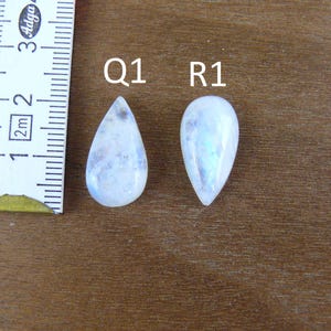 May include: Two pear-shaped, translucent white moonstones labeled Q1 and R1, positioned next to a white measuring tape showing a scale in centimeters. The stones exhibit a smooth, polished surface with subtle internal inclusions and blue sheen.