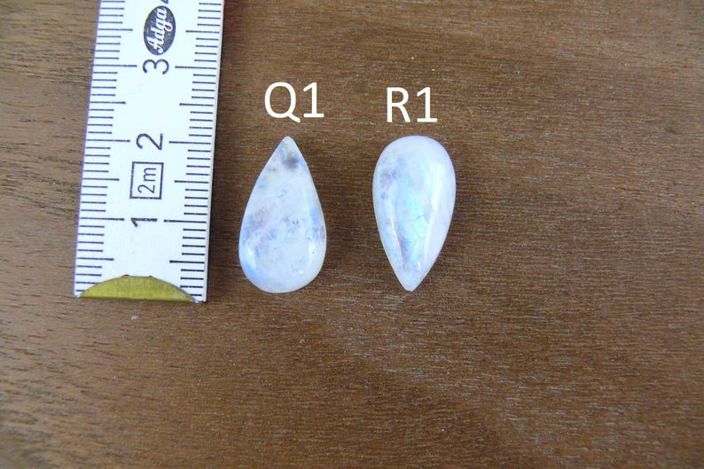 May include: Two pear-shaped, translucent white moonstones labeled Q1 and R1, positioned next to a white measuring tape showing a scale in centimeters. The stones exhibit a smooth, polished surface with subtle internal inclusions and blue sheen.