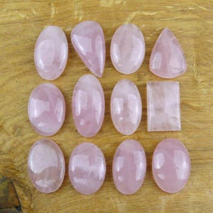 Puede incluir: Una colección de doce piedras preciosas de cuarzo rosa pulidas dispuestas en una cuadrícula. Las piedras presentan varias formas, incluyendo óvalos, una lágrima y un rectángulo. Cada pieza muestra una superficie rosa pálido, lisa y translúcida.