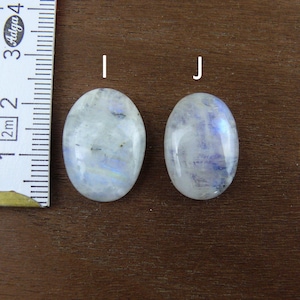 Puede incluir: Dos piedras preciosas de forma ovalada, de piedra lunar, con matices iridiscentes azules y blancos. Se coloca una regla junto a las piedras para medir. Las piedras est&aacute;n sobre una superficie de madera marr&oacute;n.