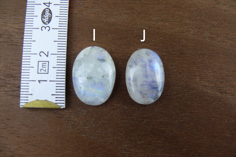 Puede incluir: Dos piedras preciosas de forma ovalada, de piedra lunar, con matices iridiscentes azules y blancos. Se coloca una regla junto a las piedras para medir. Las piedras est&aacute;n sobre una superficie de madera marr&oacute;n.