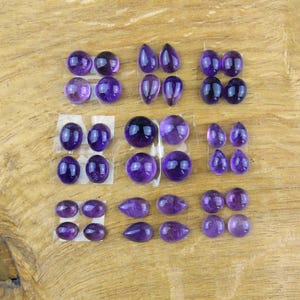 Könnte beinhalten: Eine Sammlung polierter Amethyst-Edelsteine in verschiedenen Formen, darunter oval, tropfenförmig und rund. Die Edelsteine haben eine tiefviolette Farbe und sind auf einer Holzoberfläche angeordnet.