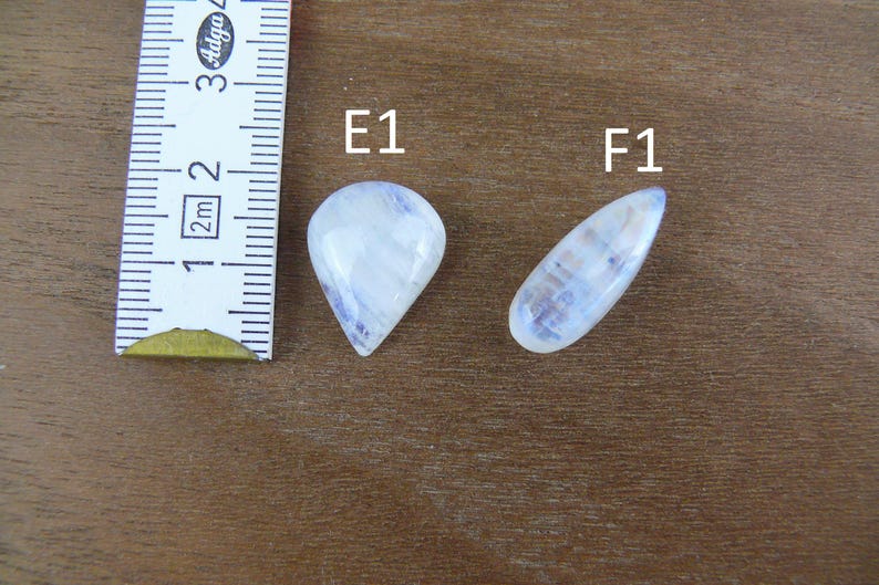 May include: Two polished moonstone cabochons, labeled E1 and F1, are displayed next to a ruler showing measurements in centimeters. The E1 stone is pear-shaped, while the F1 stone is elongated and teardrop-shaped, both featuring a translucent white appearance.