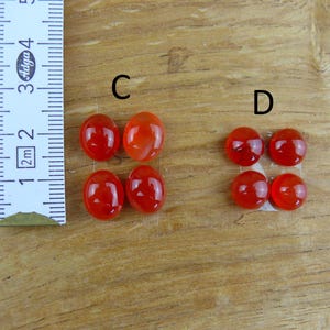 Op de afbeelding: Een verzameling rood-oranje edelstenen, sommige ovaal en sommige rond, liggen op een houten oppervlak. Een liniaal is geplaatst voor schaal. De edelstenen hebben een diameter van ongeveer 2.5 cm.