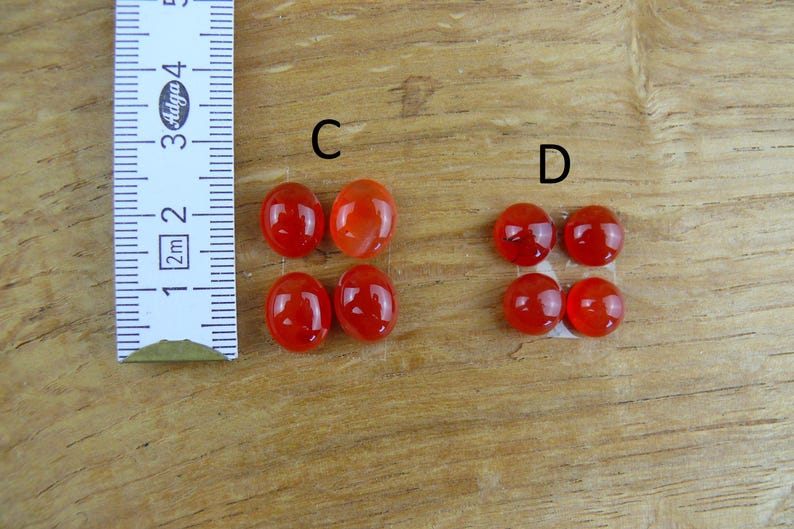 Op de afbeelding: Een verzameling rood-oranje edelstenen, sommige ovaal en sommige rond, liggen op een houten oppervlak. Een liniaal is geplaatst voor schaal. De edelstenen hebben een diameter van ongeveer 2.5 cm.