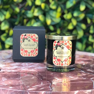 Puede incluir: Un tarro de vela de vidrio negro con tapa y etiqueta doradas. La etiqueta dice "Punjab Da Gulab Tuberose & Orange Blossom #experiencepakistan". La vela está sobre una superficie de mármol.