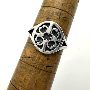 May include: A silver ring featuring a circular face with an intricate, openwork cutout design. The center of the ring displays a dark, recessed background behind the polished silver metalwork, which forms a symmetrical pattern of curved shapes.