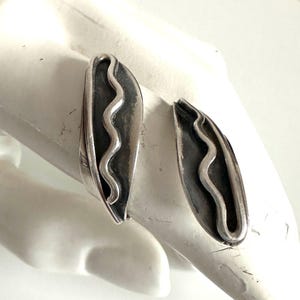 May include: A pair of silver-toned earrings. Each earring has a leaf-like shape with a wavy, textured design in the center. The earrings are displayed on a white surface, showcasing their metallic sheen and intricate details.