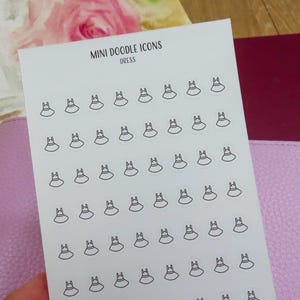 May include: A white card with the text "MINI DOODLE ICONS DRESS" and small dress icons. The card is held over a pink leather notebook and a burgundy notebook. A watercolor painting of flowers and a cup is in the background.