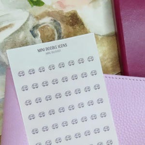 Könnte beinhalten: Ein Stickerbogen mit Mini-Doodle-Icons von Autos, beschriftet mit "Mini Doodle Icons". Die Aufkleber liegen auf einer hellrosa, ledertexturierten Oberfläche, mit einem weinroten Notizbuch und einem Aquarell im Hintergrund.