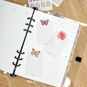 K&ouml;nnte beinhalten: Drei transparente Plastik-Seitenteiler mit Blumen- und Schmetterlingsmotiven. Die Teiler sind mit "MONAT", "SO" und "SA" beschriftet.