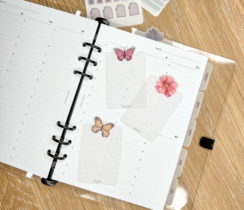 K&ouml;nnte beinhalten: Drei transparente Plastik-Seitenteiler mit Blumen- und Schmetterlingsmotiven. Die Teiler sind mit "MONAT", "SO" und "SA" beschriftet.