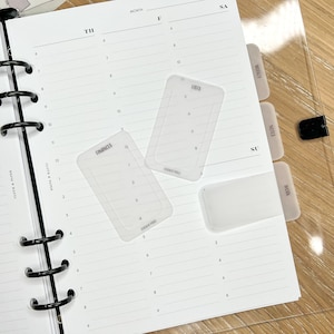 K&ouml;nnte beinhalten: Ein wei&szlig;er Planer mit schwarzen Ringen und einer transparenten Plastikh&uuml;lle. Der Planer hat Registerkarten mit den Beschriftungen "W&ouml;chentlich", "Notizen", "SO" und "Arbeit". Es gibt drei transparente Plastikkarten mit Zahlen darauf, beschriftet mit "FINANCES", "10/13" und "STICKY PRO".