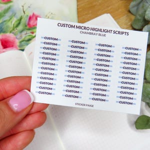 May include: A white sticker sheet with the text "CUSTOM MICRO HIGHLIGHT SCRIPTS CHAMBRAY BLUE". The sheet has multiple rows of the word "CUSTOM" in a blue font. The sticker is held over an open notebook with floral illustrations.