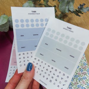 Puede incluir: Hojas de pegatinas blancas con texto e iconos. Una hoja está etiquetada como "TABS CHAMBRAY BLUE" y la otra como "TABS SPROUT GREEN". Cada hoja tiene secciones para meses, diario, notas e índice.