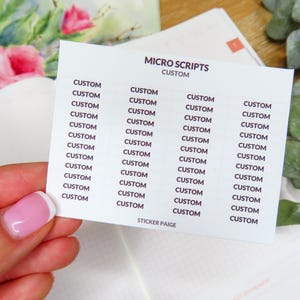May include: A white sticker sheet with the text "MICRO SCRIPTS CUSTOM" printed in black. The sheet is held by a hand with a pink and white manicure. The background includes a floral design and greenery.