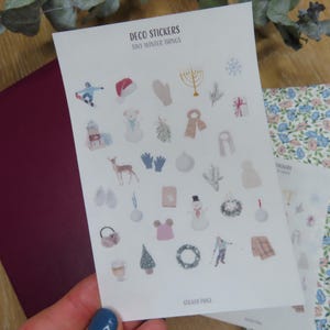 May include: A sheet of Deco Stickers with Tiny Winter Things. The stickers feature winter-themed illustrations, including a snowman, mittens, a snowflake, and a deer. The sheet is held over a burgundy envelope and a floral patterned notebook.