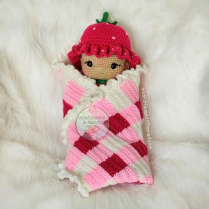May include: A crocheted strawberry doll wrapped in a pink and white checkered blanket. The doll has a red hat with green leaves and a pink and white crocheted dress. The blanket has a crocheted border.