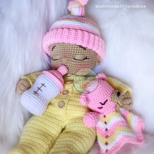 May include: A crocheted baby doll with closed eyes, wearing a yellow and white striped outfit, a pink hat, and holding a white bottle. The doll is holding a pink crocheted teddy bear wearing a pink, white and yellow striped blanket.