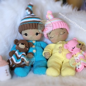 May include: Two crocheted baby dolls, one in blue pyjamas with a brown teddy bear and the other in yellow pyjamas with a pink teddy bear. Both dolls have brown yarn hair and are wearing crocheted hats.