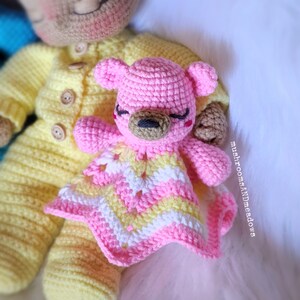 May include: A pink crocheted teddy bear with a yellow and pink blanket. The bear has a brown nose and closed eyes. The bear is lying on a white surface.