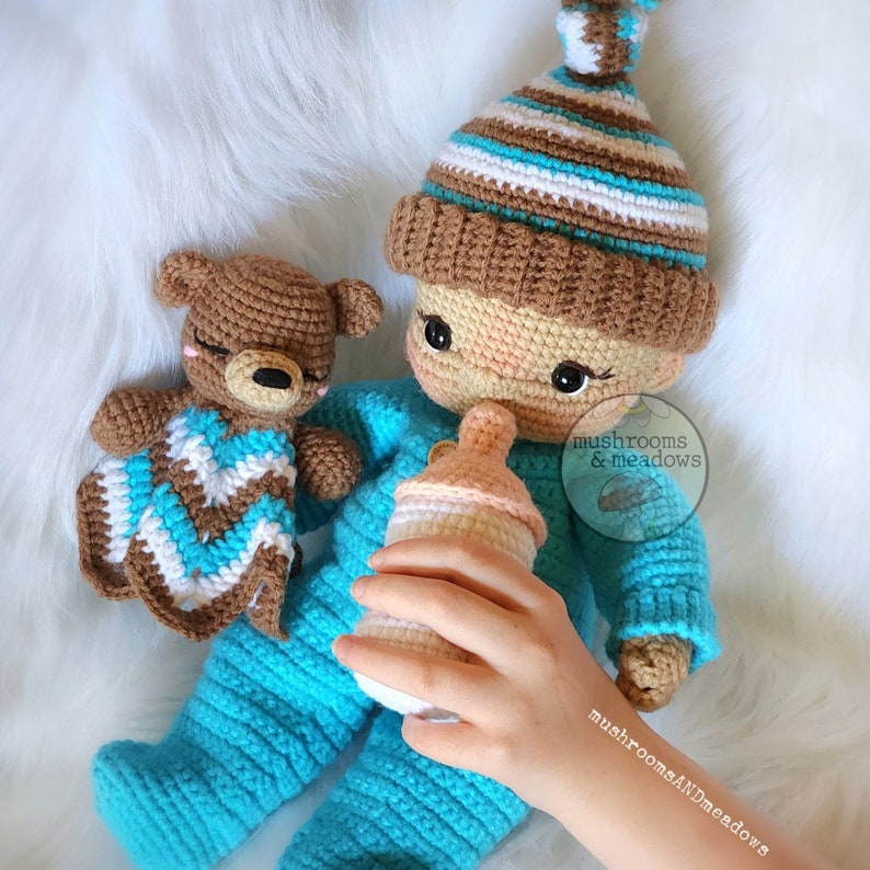 May include: A crocheted doll wearing a blue and white striped hat and a blue outfit, holding a bottle. A brown crocheted bear with a white and blue blanket is in the background. The doll is labelled "mushrooms & meadows".
