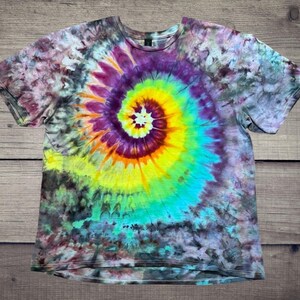 May include: A short-sleeved tie-dye t-shirt with a spiral design. The spiral starts with a white center, transitioning through yellow, orange, purple, and blue hues. The shirt's background has a mix of dark purple, gray, and teal colors.